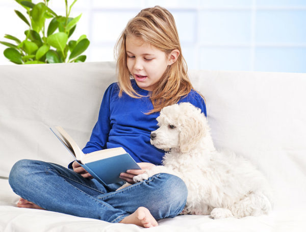 blond young girl reading a book to her poodle puppy on the couch