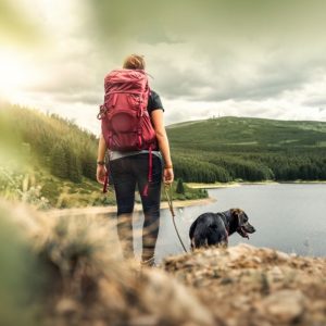 Best dog breeds for hiking and outdoors Hiker wearing a red backpack stands with back to camera looking over a lake, with a black dog alongside